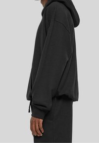 Persona vestida con un sudadera negra holgada y pantalones negros, de pie contra un fondo gris liso, en vista lateral.