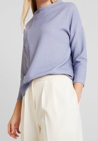 Ljuslila, strukturerad stickad tröja med hög hals, trekvartsärmar och avslappnad passform, i kombination med krämfärgade vida byxor.