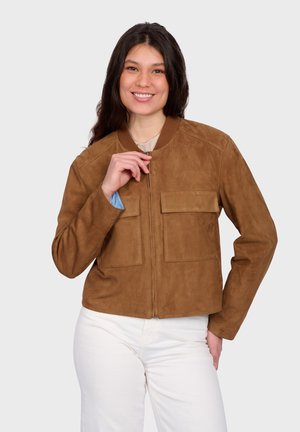 Lächelnde Frau schließt eine braune Wildlederjacke mit Fronttaschen, trägt weiße Hosen und ein helles Hemd vor einem einfarbigen Hintergrund.