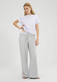 Vrouw in een wit shirt met korte mouwen en lichtgrijze wijde sweatpants, die met één hand haar shirt iets oplicht, blootsvoets op een witte achtergrond.