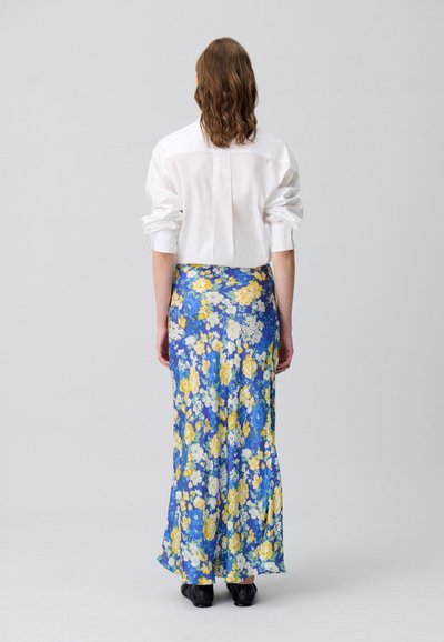 Jupe longue à fleurs bleue avec des fleurs jaunes et blanches, associée à une chemise blanche à boutons. Tissu lisse, longueur chevilles, volant subtil à l'ourlet.