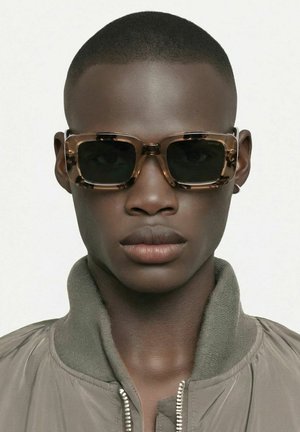 Homme aux cheveux courts portant des lunettes de soleil carrées en écaille et une veste grise zippée, sur un fond clair uni.