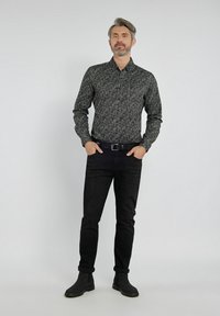 Czarna, wzorzysta koszula na guziki o slim fit, połączona z ciemnymi dżinsami i czarnymi botkami za kostkę. Mężczyzna stoi pewnie, z rękami w kieszeniach.