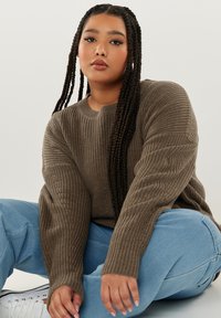 Brauner gerippter Pullover mit langen Ärmeln, rundem Ausschnitt und strukturiertem Stoff, kombiniert mit hellblauen Hosen und weißen Turnschuhen.