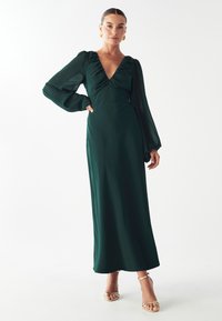 Dlhé zelené šaty s hlbokým V-výstrihom a nariasenou sukňou. Majú priesvitné rukávy a splývavú sukňu. Skombinované so zlatými sandálmi na vysokom opätku.