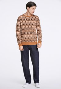 Strickpullover mit einem braunen, orangefarbenen und cremefarbenen Muster, das Tiere zeigt, kombiniert mit dunklen Jeans und weißen Turnschuhen. Gerippte Bündchen und Saum.