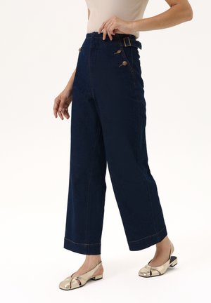 Frau trägt dunkelblaue, hoch taillierte, weit geschnittene Jeans mit Knopfdetails und beige Slingback-Schuhe mit niedrigem Absatz.