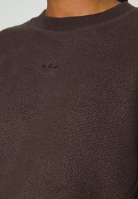 Brązowy sweter z polaru z teksturowaną powierzchnią oraz ciemnobrązowym haftowanym logo Adidas na piersi. Żebrowany kołnierz dla lepszego dopasowania.