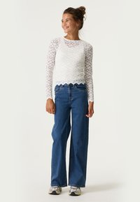 Weißes Spitzenshirt mit langen Ärmeln und gewelltem Saum, kombiniert mit hoch taillierten blauen Schlagjeans und grauen Sneakers. Glatte, strukturierte Materialien.