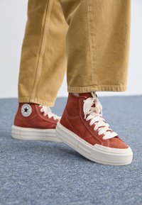 Zapatillas altas de ante rojo con cordones blancos, superficie texturizada y suela de goma blanca gruesa. Presenta un logo circular en el costado.