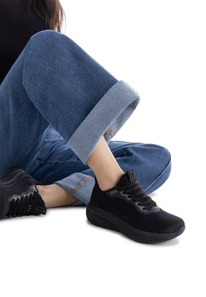 Zapatilla deportiva negra hecha de malla y caucho, con un upper texturizado, suela gruesa y cordones negros, combinada con pantalones de mezclilla.