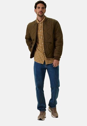 Veste bomber matelassée vert olive sur une chemise à carreaux marron et jaune, jean bleu et baskets beiges. Style décontracté, coupe relax.