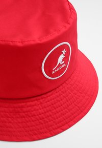 Cappello a secchiello rosso realizzato in nylon, con un logo Kangol ricamato in bianco all'interno di un contorno circolare. Texture liscia e visiera ampia.