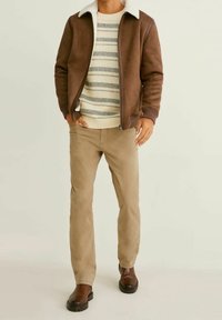 Uomo che indossa un giaccone in shearling marrone, un maglione a righe color crema, pantaloni beige e stivali di pelle marroni, in piedi su uno sfondo chiaro.