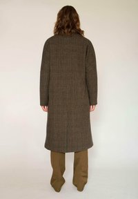 Manteau à carreaux marron avec une coupe longue et droite. Fabriqué en tissu texturé. Porté avec un pantalon kaki, présenté de dos.