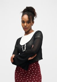 Jeune femme aux cheveux bouclés portés en chignon haut, portant un cardigan noir transparent par-dessus un haut blanc et une jupe bordeaux avec des motifs de cœurs blancs, bras croisés.