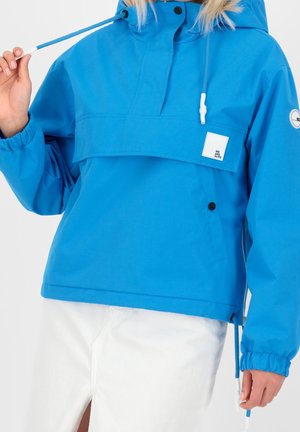 Persona que lleva una chaqueta con capucha de color azul brillante con bolsillo frontal con solapa y pantalones blancos, ajustando el cordón azul de la capucha de la chaqueta.