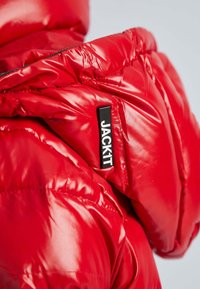 Rode pufferjack met glanzende afwerking, voorzien van een hoge kraag en zwart logo-label. De stof is gevoerd voor isolatie.