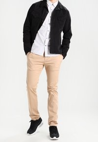 Black jacket with two chest pockets, white button-up shirt with small black dots, beige pants, and black sneakers.