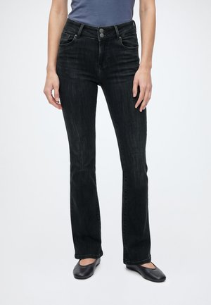 Femme portant un jean bootcut noir taille haute associé à des chaussures plates noires, debout devant un fond clair uni.