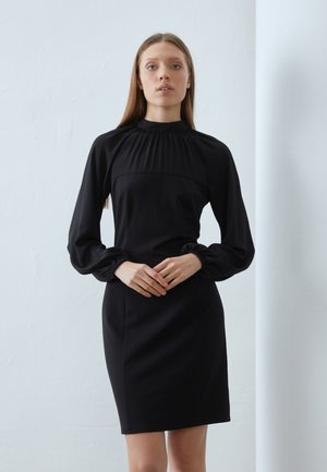 Svart kjole med lange, puffede ermer og høy hals med samlet detaljer. Stoffet virker glatt og strukturert, med en tettsittende silhuett.
