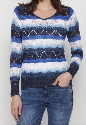 Femme portant un pull à manches longues en maille avec des rayures en zigzag bleu, blanc et marine, et un jean en denim bleu, une main dans la poche.
