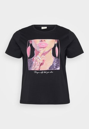 Melnā T-kreklā ar sekvīnu grafiku, kas attēlo sievietes seju ar rozā lūpām un auskariem, un tekstu "Izveido sevi, kuru mīli" zem attēla.