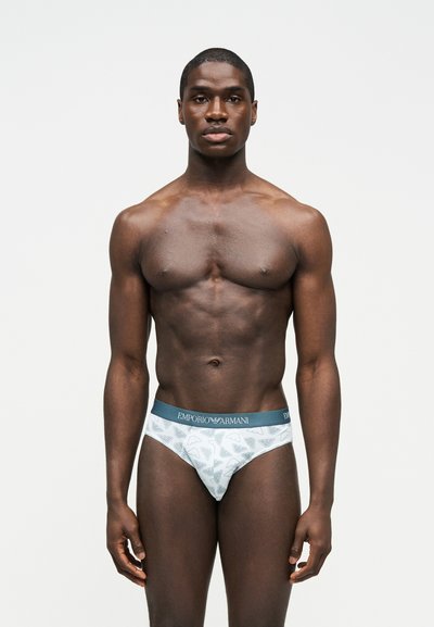 Μυώδης άνδρας στέκεται προς τα εμπρός, φοράει λευκά εσώρουχα Emporio Armani με μπλε ζώνη μέσης και μοτίβο λογότυπου σε απλό ανοιχτό φόντο.