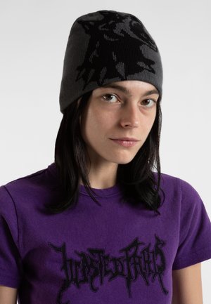 Young woman with dark hair wearing a gray patterned knit beanie and a purple t-shirt with black graffiti-style text.