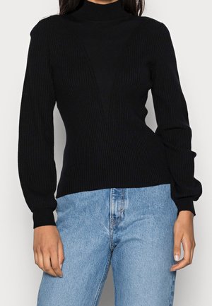 Kvinde iført sort ribbet rullekravebluse med lange ærmer og lyseblå højtaljede jeans, stående med afslappede hænder.