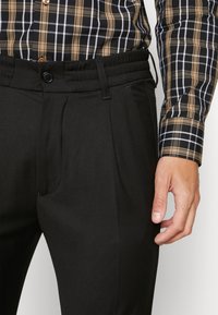 Zwarte broek met een gladde textuur, voorzien van een rechte voorkant, zijzakken en een elastische tailleband. Gecombineerd met een geruit overhemd met knopen.