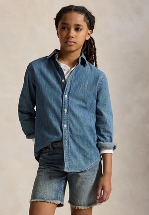 Nuori henkilö, jolla on letti hiukset, pukeutuneena siniseen farkkupaitaan valkoisen paidan päällä ja rispaantuneisiin farkkushortseihin, seisoo neutraalia taustaa vasten.