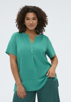 Kobieta o kręconych włosach, ubrana w zieloną bluzkę z krótkim rękawem i guzikami oraz pasujące zielone spodnie, pozująca na tle jednolitego jasnego tła.