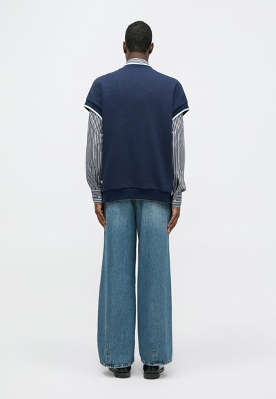 Sweatshirt militar oversized com mangas longas listradas, combinado com calças de ganga largas. Textura suave, design casual e cores contrastantes.