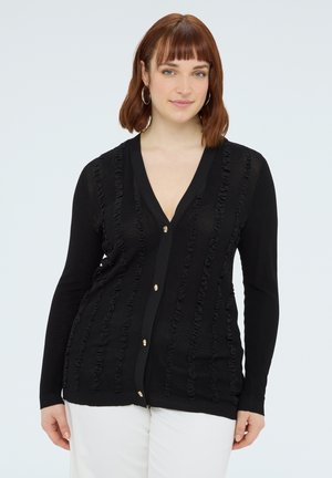 Młoda kobieta o rudych włosach, ubrana w czarny, teksturowany kardigan na guziki oraz białe spodnie, stojąca na tle jasnego, jednolitego tła.