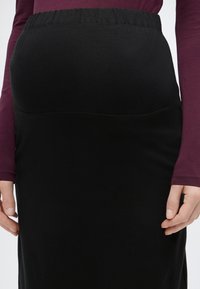 Kobieta ubrana w czarną ołówkową spódnicę z elastycznym pasem i długi rękaw ciemnofioletowej bluzki, ręce swobodnie opuszczone wzdłuż ciała.