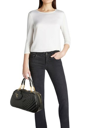 Borsa a mano in pelle nera con motivo a cuore trapuntato, dettagli dorati, tenuta in mano. Modella indossa un top bianco e jeans scuri.