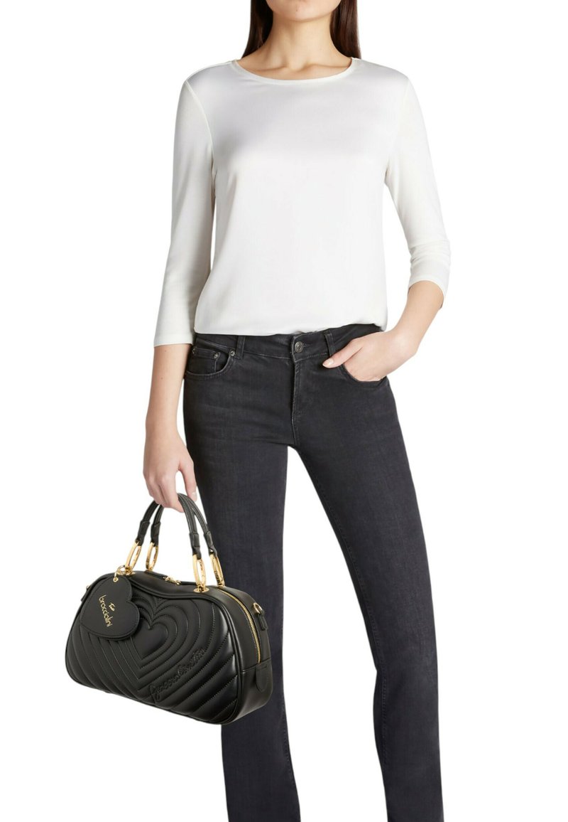 Sac à main en cuir noir avec motif matelassé en forme de cœur, accents dorés, tenu à la main. Le mannequin porte un haut blanc et un jean foncé.