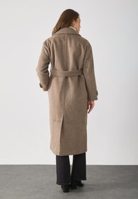 Lange beige wollen jas, met een ceintuur in de taille, een brede kraag en zijsplitten. Gedragen over zwarte flared broeken en laarzen met hak.