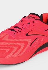 Φωτεινό κόκκινο παπούτσι για τρέξιμο Reebok με μαύρες λεπτομέρειες, λεπτομερές πλέγμα στο επάνω μέρος και κόκκινα κορδόνια που ταιριάζουν, σε λευκό φόντο.