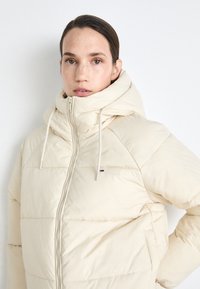 Cream puffer jacket with a high collar, front zipper, and drawstring, featuring quilted texture and subtle logo accent.