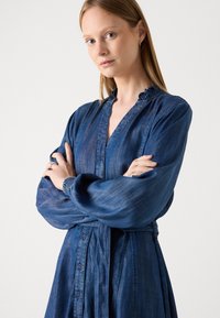 Temno modra, dolga obleka z rokavi, z nabrano ovratnikom, zapenjanjem na gumb in zbranim manšetami. Oblačilo je z pasom in ima tekočo krilo.