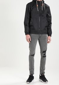 Zwarte waterdichte jas met een rits aan de voorkant, twee borstzakken en elastische boorden. Gecombineerd met gescheurde grijze jeans en zwarte sneakers.