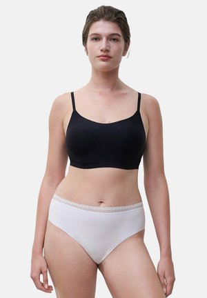 Woman standing facing forward wearing a black sleeveless crop top and white high-waisted underwear with lace trim on a plain background.