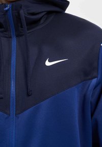 Tums zips, tumši zila krāsā, izgatavota no gludas, vieglas auduma. Iezīmēts ar izteiksmīgu baltu Nike swoosh uz krūtīm un ar auklām kapucei.