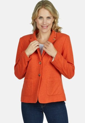 Lachende vrouw die een feloranje blazer met twee knopen en voorzakken rechtzet, draagt een donkerblauwe spijkerbroek tegen een effen achtergrond.