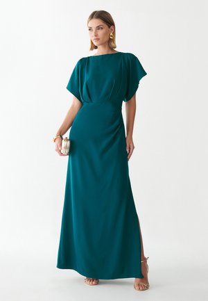 Vrouw met een teal-kleurige, vloerlange jurk met korte mouwen, die een kleine gouden clutch vasthoudt en gouden oorbellen en sandalen draagt.
