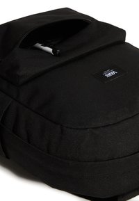 Svart ryggsäck med texturerat tyg, ficka med dragkedja upptill och en logotypmärke. Har justerbara remmar och en kompakt, strömlinjeformad design.