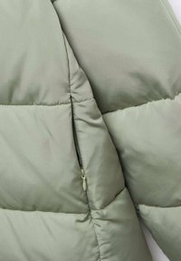 Steppjacke in hellem Olivgrün mit glatter Textur, ausgestattet mit einer Reißverschlusstasche und gesteppter Naht für Isolierung.
