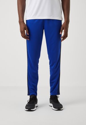 Man wearing blue athletic pants with black side stripes and black running shoes, standing against a plain white background.